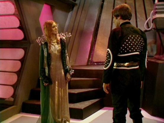 Claude 3.7 describes the image as: "This image appears to be from a science fiction television series, featuring a scene set inside what looks like a futuristic spacecraft or space station. The interior has distinctive angular architecture with pink/reddish lighting elements and metallic walls.

Two figures are shown in conversation. On the left is a person wearing an elaborate costume consisting of a golden/beige dress with a green robe or cloak. On the right is a person dressed in black attire with decorative studded details on the back of their jacket.

The setting has a distinctive 1970s-80s sci-fi aesthetic with its dramatic lighting and geometric set design. The costumes have a theatrical quality typical of British science fiction productions of that era, combining futuristic elements with somewhat ornate styling."

Blakes7Bot, version 3.9.30.