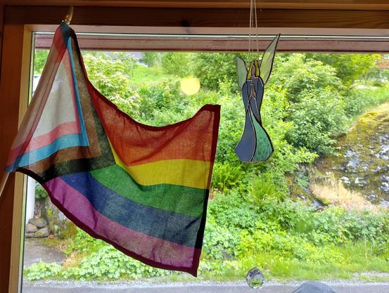 Eit vindauge der det heng eit progressive prideflagg, og ein engel med folda hender og vengjer i glaskunst med gult hår og blå kjortel. Ute er allting grønt og triveleg.