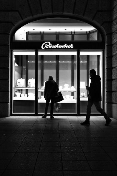 black and white. portrait format. Shot in the early evening. It is already dark. City scene on a shopping street. The stage/background is a brightly lit shop window of a jewelry store. A woman stands in front of it and looks at the goods. A man walks past the scene and looks at the woman again. Both appear as a dark silhouette.