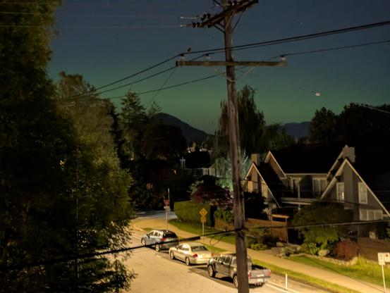 A night scene looking north towards the small mountains silhouetted against the faint greenish and pinkish aurora. A few vehicles are parked on the street below. Some houses are across the street. Lots of trees are in the picture. A wooden utility pole festooned with various wires is front and centre.