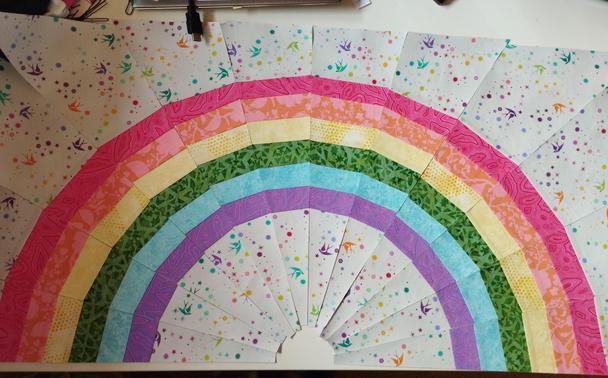 A photo of a paper pieced rainbow laid out before the final sewing of all pieces. The colors of the rainbow are desaturated and the background fabric is grey with stars and swallows on it in all colors of the rainbow.