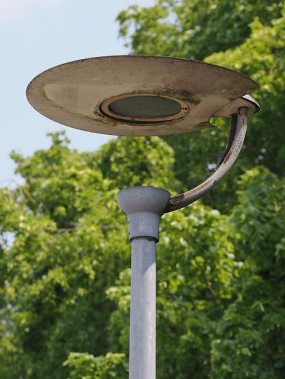 Eine Straßenlaterne, bestehend aus einer flachen Scheibe, die mittels eines runden Trägers frei schwebend über dem Lampenpfosten befestigt ist. In der Mitte der Scheibe leuchtet eine runde Fläche senkrecht nach unten. Im Hintergrund sind unscharf von der Sonne beschienene Bäume zu sehen.