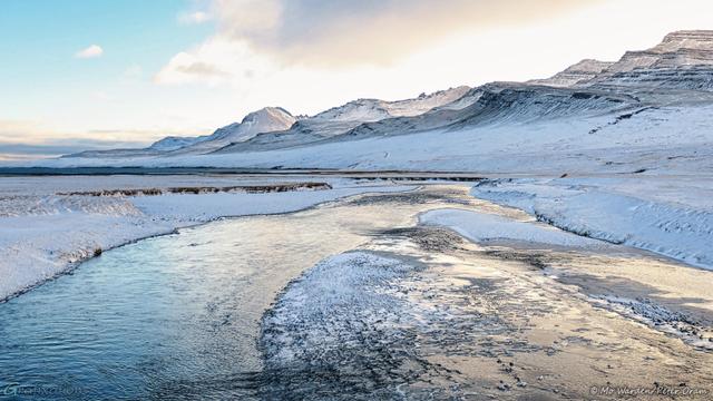 A colour photo of a landscape with a river running away from the viewpoint and towards a body of water in the distance. The ground is covered in snow and ice. The sky is cloudy with clear cyan patches, and a range of mountains is on the horizon. The sky colours are reflected in the water surface.