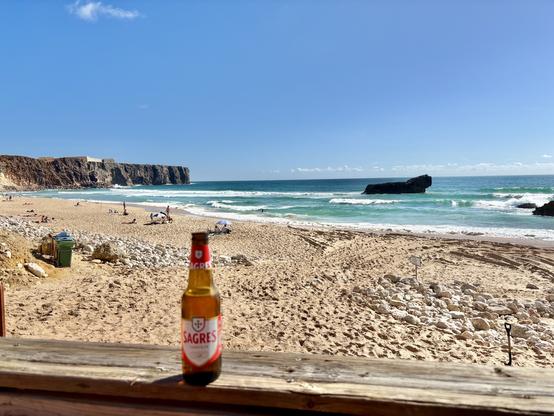 A bottle of Sagres beer sits on a wooden surface in the foreground, overlooking Tonel beach in Sagres, Portugal with scattered people relaxing. The ocean waves are visible in the background, along with cliffs and a rock formation in the water under a clear blue sky.