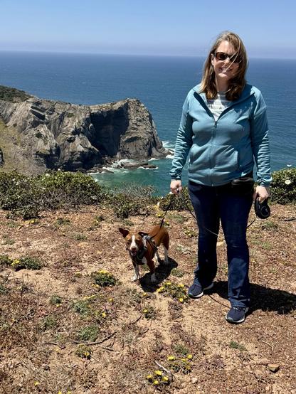 A woman stands on a rocky coastal trail with a smiling dog beside her. The background features a dramatic ocean view and rugged cliffs under a clear blue sky. Wildflowers grow on the ground around them.