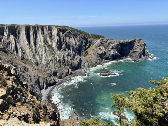 A scenic view of rugged coastal cliffs meeting a turquoise ocean. The cliffs have distinct vertical striations, and patches of greenery are visible along the edges. The sky is clear with a few wispy clouds, enhancing the natural beauty of the landscape.