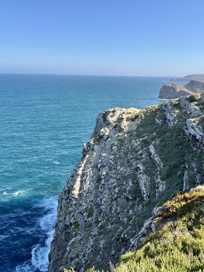 A coastal landscape featuring a rocky cliff overlooking a vibrant blue ocean under a clear sky. The scene includes rugged terrain with patches of greenery, highlighting the natural beauty of the coastline.