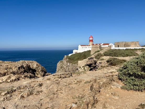 A coastal landscape featuring a lighthouse with a red dome, situated on a rocky outcrop overlooking the ocean. Nearby, there is a white building and remnants of a stone fortification. The sky is clear and blue, enhancing the view of the sea.