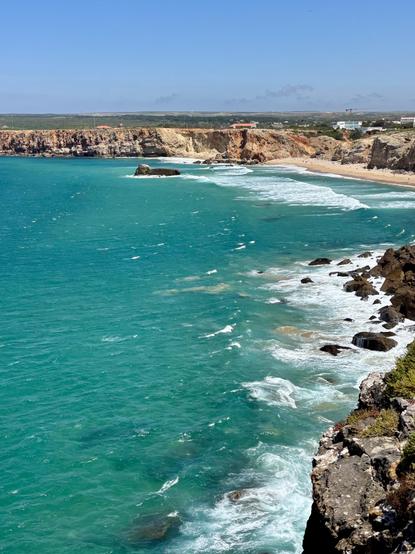 A vibrant coastal scene featuring turquoise waters and waves crashing against rocky shores. Cliffs line the beach, and a sandy area is visible in the background with a few buildings. The sky is clear and blue, adding to the picturesque landscape.