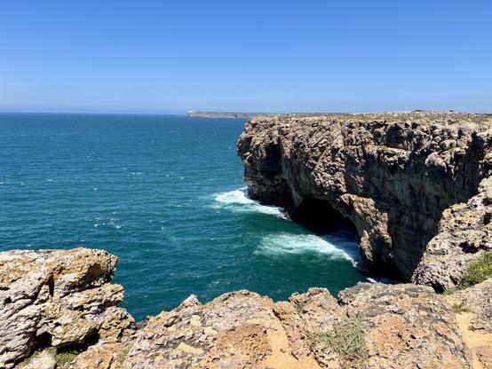 A rocky coastline meets the deep blue ocean under a clear sky. Waves crash against the cliffs, creating a scenic contrast between the rugged rock formations and the tranquil water. The image captures a serene and natural coastal landscape.
