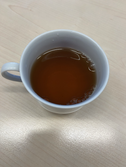Eine weiße Tasse mit mahagonifarbenem Tee darin auf einer hellen Tischplatte. Licht spiegelt sich am Rand der Tasse und in einigen Luftblasen.
