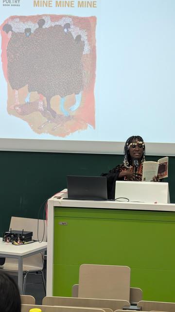 Uhuru Portia Phalafala holding her book "Mine Mine Mine", in the process of reading from it. The background shows an enlarged picture of her book cover
