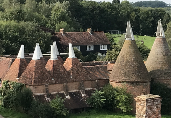 An elevated view of oasthouses. On the left a, 2x3 group of six oasts, each built on a square plan and topped by their characteristic cowls. On the right are two larger oasts built to a circular plan. The buildings are brick built with tiled conical roofs. Behind the oasts is a residential building, black walled and tiled and surrounded by trees. The sun is shining.