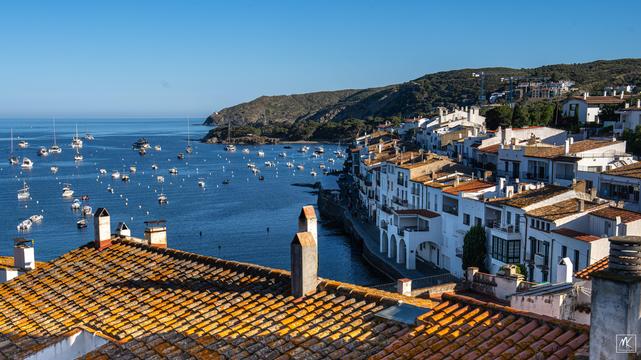 Color photo of a seaside town of white washed buildings on the Spanish Costa Brava, with many small boats in the water offshore.