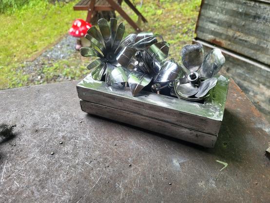 Une petite boite en métal est déposée sur une table de travail extérieur. 3 fleurs trônent sur le dessus.