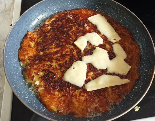 In einer Bratpfanne, bildfüllend, ist ein Rösti, eine Art Pfannkuchen aus grib geriebenen Kartoffeln, teilweise mit Käse überbacken.