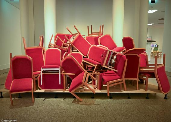 In einem Museum wurden auf einer kleinen Fläche dutzende rot gepolsterte Stühle chaotisch übereinander gestapelt. /
In a museum, dozens of red upholstered chairs were chaotically stacked on top of each other in a small area.