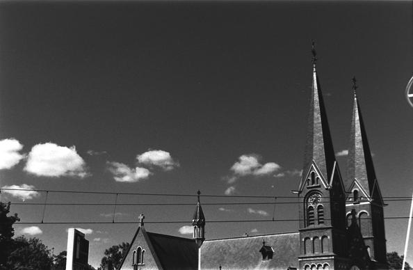 Black and white photograph, church roof and two bell towers against dark clear sky, with small white clouds.