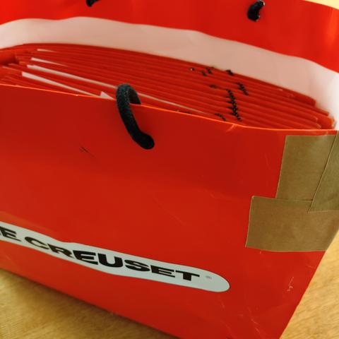 foto einer orangenen tragetasche aus festem papier mit dem schriftzug der firma "le creuset", darin orangefarbene sammelmappen