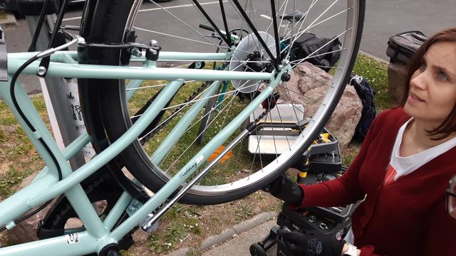 Une jeune femme vérifie le résultat de son travail de dévoilage de roue arrière de vélo, posé sur un pied d'atelier pîle au-dessus d'une servante à outils -- sur un large trottoir piétons.