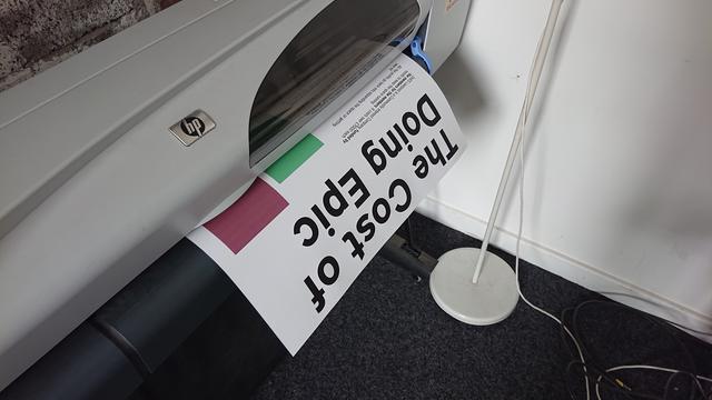 Our 42in large format printer in the middle of printing a poster. The title is visible, reading "The cost of Doing Epic", and some text and bar chart are just becoming starting to show as it leaves the printer.

There's a red bar and a green bar, but the green bar doesn't go as high up as the red one...