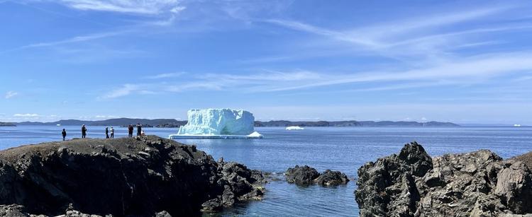 A rugged angular coastal landscape with six people viewing a blocky iceberg grounded near shore. Thin wispy clouds crisscross a blue sky above an ocean with similar shades of blue. A large iceberg in the mid ground holds the attention of the onshore onlookers, with several small icebergs in view in the distance. The large iceberg itself has an arch near the bottom and darker horizontal striatuins near the top , with shades of lighter greenish blue. [Twillingate (NL, Canada) May 2025]