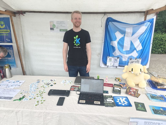 Tobias Fella, a tall, blond German contributor wearing a KDE Eco t-shirt, stands behind the table at the KDE booth at the Umwelfestival in Berlin. On the table you can see stickers, phones, a Steam Deck, computers, all running KDE software of one kind or another... And, oh yeah, a cute Mastodon plushy.