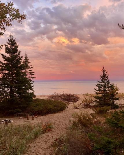 A scenic view of a sandy beach path on Beaver Island leading to a tranquil Lake Michigan, framed by evergreen trees. The sky is filled with vibrant pink and orange hues as the sun sets, reflecting softly on the water's surface.