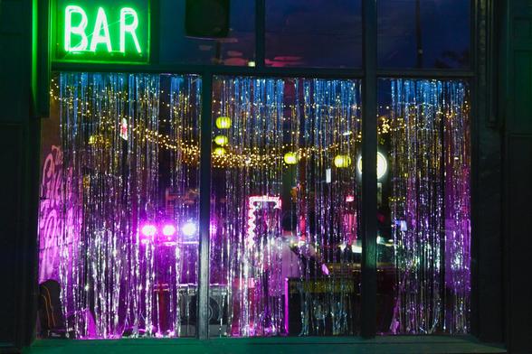 This photo shows the front window of a lively bar at night. A neon green "BAR" sign glows in the top left corner, casting light onto the dark frame. Behind the large window, long, shimmering silver tinsel hangs from top to bottom, partially obscuring the view inside. Through the tinsel, bright purple, pink, and yellow lights fill the room with a festive, almost dreamlike glow. Warm string lights and round paper lanterns hang across the ceiling. Silhouettes of people and bar equipment are faintly visible, creating a sense of motion and celebration inside this colourful, inviting nightlife scene.