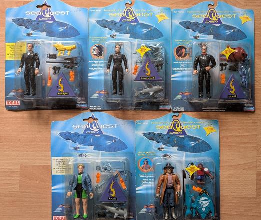 5 seaQuest-Actionfiguren in zwei Reihen auf Fußboden: Oben links Bridger, iMitte Hitchcock, rechts O'Neill. Unten links Lucas, rechts der Regulator. Zu jeder Actionfigur ist Zubehör in Form futuristischer Waffen gepackt, manchmal mit direktem Bezug zur Figur.