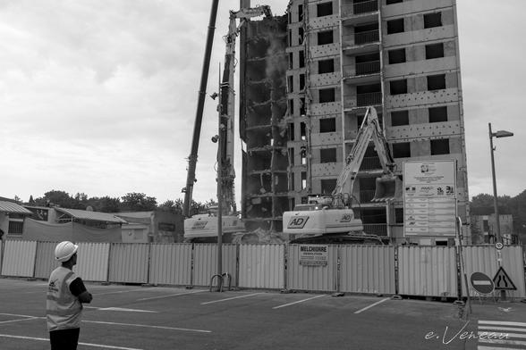 A man wearing a hard hat looks out over a demolition site where two pieces of mechanical equipment are attacking a ten-storey building.