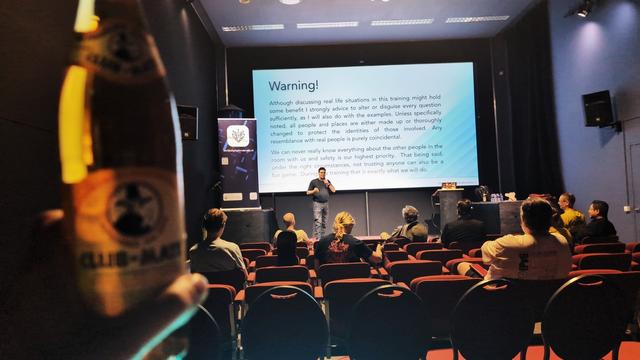 Still empty conference room (about to become more busy) with Marcel on stage with behind him a giant warning to not discuss real cases blindly because you don't know who else is listening. The photographer is in the back of the room holding a Club Mate drink in the picture.