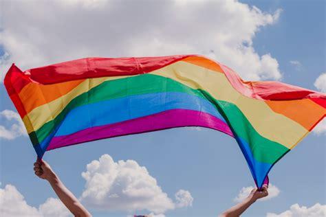 A person is holding up a large rainbow Pride flag in the air against a bright blue sky with fluffy white clouds. The flag’s colors are vibrant and ripple in the wind, symbolizing LGBTQ+ Pride and the importance of visibility and celebration.