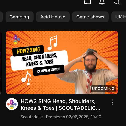 A screenshot of a thumbnail from a video sharing app video. It has a Scout Leaders with a red and lint grey necker around their neck with his hands touching his head at hair level. 

Alongside him is the wording How2 Sing Head Shoulders Knees And Toes, Campfire Songs. 

The thumbnail also has wording saying Upcoming, a date for what is the next day and time of day in the morning or 10am.

It is to be released on the Scoutadelic Channel.