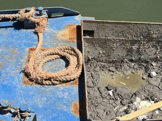 Colour photo of part of a construction boat. The left half of the frame is blue metal deck, stained and rusted and corroded, with a thick rusty-coloured rope coiled up in a circle, and one frayed end knotted to the edge of the boat. The right half of the frame is the boat's container for rubbish, which is caked with grey-brown dried cracked mud, and there is a thin plank of blond wood lying diagonally across the bottom right hand corner. There is a thin strip of green canal water along the top of the frame. The feeling of the image is one of tools having been emphatically downed, and the equipment left to very much take care of itself while its operators are off having a siesta.