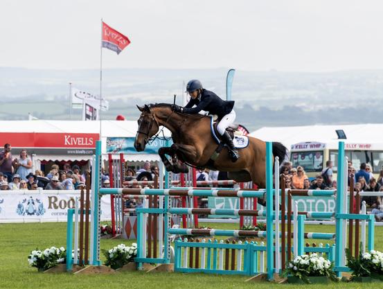 A female rider and her large bay horse clearing a double fence on a showjumping course