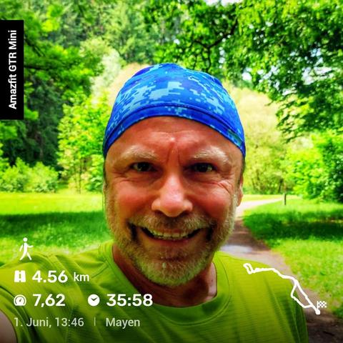 Eine Person macht ein Selfie in einem sonnigen Park. Sie trägt ein hellgrünes T-Shirt und ein blaues Kopftuch mit Tarnmuster. Im Hintergrund sind grüne Bäume, eine Wiese und ein schmaler Weg zu sehen.