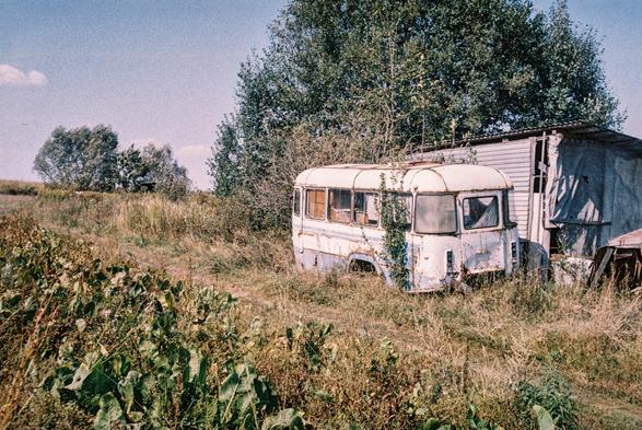 The image depicts a rural, overgrown field with an old, abandoned bus sitting beside a small makeshift structure. The bus is white with patches of rust and wear, indicating many years of neglect. Vegetation has started to overtake the vehicle—vines are growing up its side, and tall grass surrounds it. Behind the bus stands a dense cluster of trees, and in the distance, more trees line the horizon under a clear blue sky. The scene has a nostalgic, almost melancholic atmosphere, captured on film, with the grainy texture and faded colors.