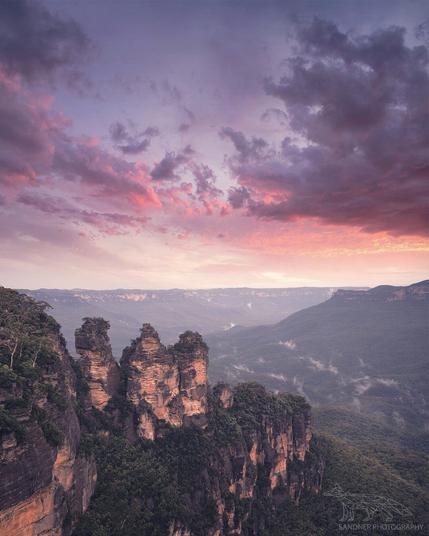 A stunning sunrise illuminates the iconic Three Sisters rock formation in the Blue Mountains, New South Wales, Australia. Warm light kisses the rugged cliffs as dramatic clouds paint the sky, with a vast, mist-filled valley stretching into the distance.