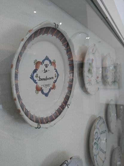 Assiettes exposées au mur. La plus proche est décorée d'une inscription "la bondance"