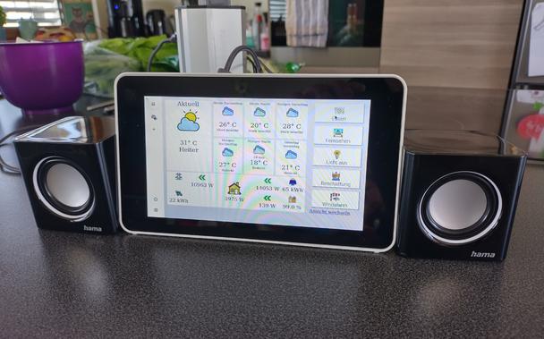 A Raspberry Pi case with Touchscreen and two small speakers to the sides. Everything is set up on a kitchen counter. The display shows a dashboard with weather forecast, energy production and use as well as some buttons to act on home automation stuff. Some cables are visible and the setup looks a bit unfinished.