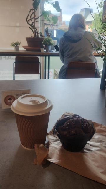 Looking past coffee and muffin on table to a person with their back to me, sitting at another table, silhouetted by light from the window