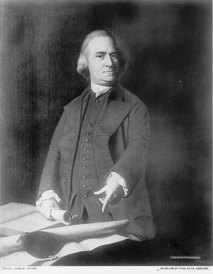 A black and white image from the Library of Congress of Samuel Adams dressed in a dark suit and pointing at a document on the table in front of him. Adams has grey hair. He is pointing with one hand and clutching a rolled up document in the other.