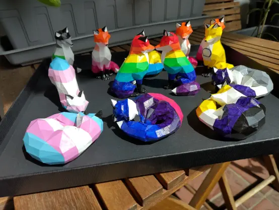 A photo of a number of 3D printed low poly foxes all painted in various pride flag colors.