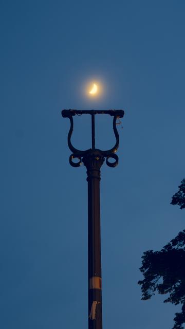 Fotografie des Mastes einer alten Straßenlaterne. Die Laterne selbst fehlt, dahinter ist allerdings ein Sichelmond zu sehen. Um den Mond herum leuchtet der nächtliche Dunst gelblich-weiß. Der Laternenmast kommt gerade von unten ins Bild und verjüngt sich nach oben. Dann verzweigt er sich in jugendstilartiger Verzierung in drei Teile, um oben in einer schmalen Plattform zu enden, auf der man sonst eine verzierte Laterne erwarten würde.