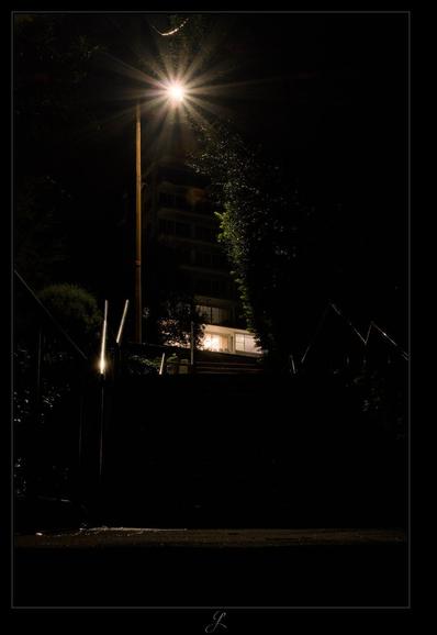 Night view from diagonally below onto some steps of a footpath with railings on the left and right. A streetlight at the end of the steps casts a strong light on the scene and shows an intense sun star. The picture is nevertheless quite dark, the railing on the left shows a reflection of the lantern light and some trees or bushes are partly lit. A high-rise building can be seen in the background, in which one apartment is brightly lit while another directly above is only dimly lit.

AI disclaimer: Using my work, its meta data, written or derived description to create media with or train AI based systems is prohibited.
