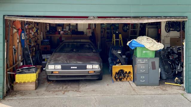 Photograph of the front of a DeLorean sports car pointed out of a crowded garage with bins, a snowblower, filing cabinets, tools, and other garage clutter visible.