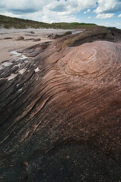 A close-up view of a large, smooth rock formation at Cheswick Sands in Northumberland, displaying striking natural patterns and swirls in shades of reddish-brown and dark grey. The rock's surface has a textured, almost layered appearance, with lines curving gracefully across it. In the background, the sandy beach stretches out towards grassy sand dunes under a partly cloudy sky, with patches of blue peeking through. The scene highlights the geological beauty and tranquil coastal environment of this Northumberland beach.