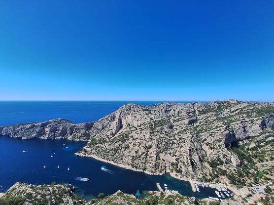 Vue panoramique d'une calanque encaissée avec un petit port de plaisance niché au creux des falaises calcaires, sous un vaste ciel bleu azur.