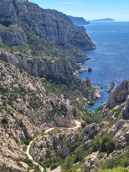 Vue plongeante sur un sentier serpentant à travers les falaises rocheuses du Massif des Calanques, menant à une crique aux eaux turquoise sous un ciel bleu.